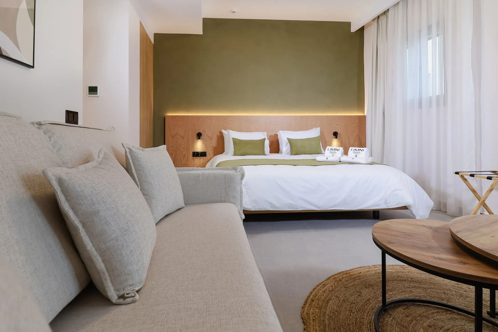 A modern hotel room with a sofa, a bed with green pillows, a round wooden table, and a headboard with integrated lighting.