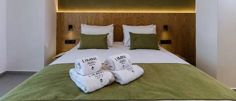 A hotel room with a wooden headboard, two white pillows, a green throw blanket, and two folded white towels with 'LIMNI' branding.