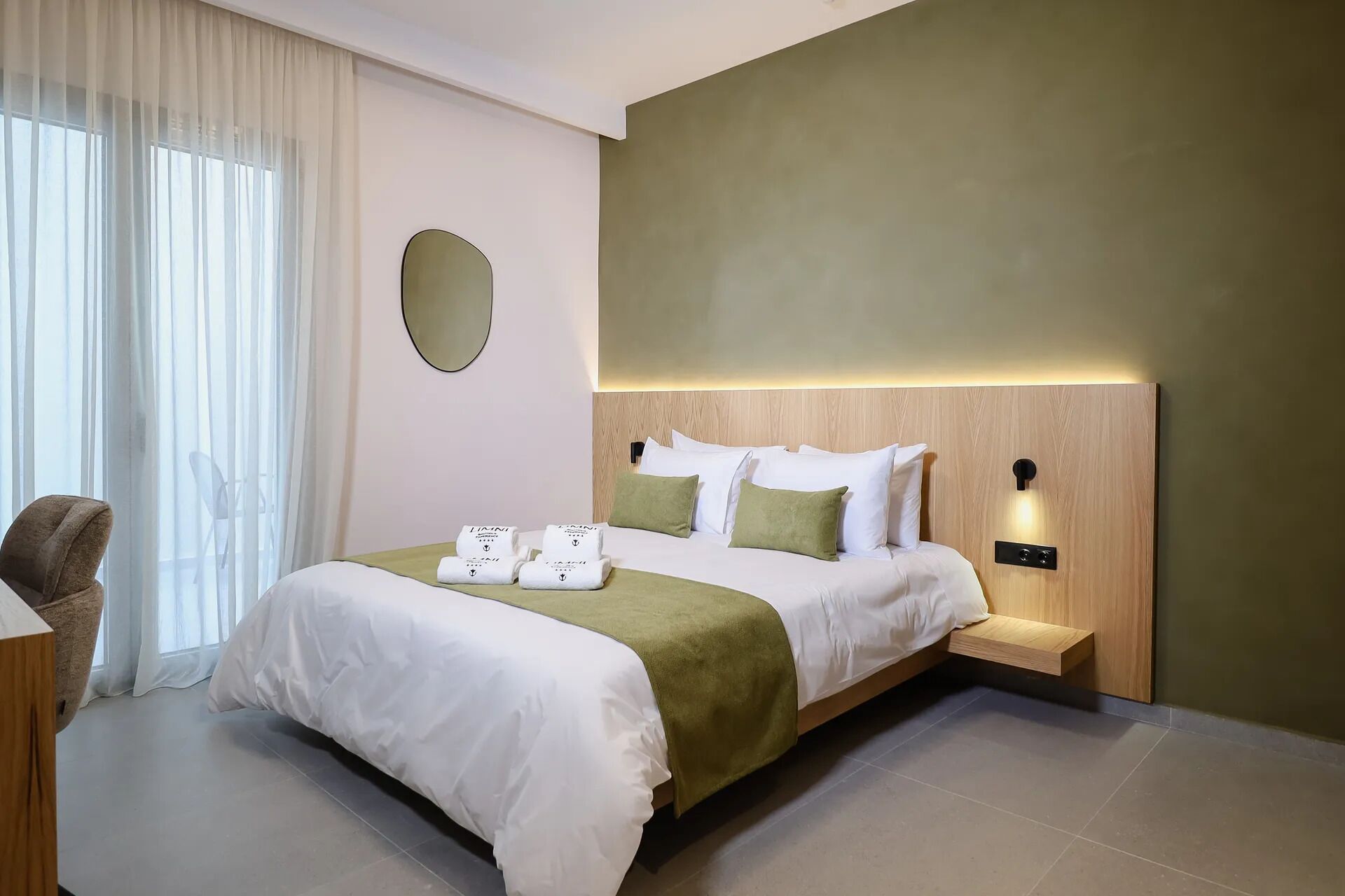 Een moderne hotelkamer met een groot bed, een nachtkastje, een stoel en een groot raam met dunne gordijnen.