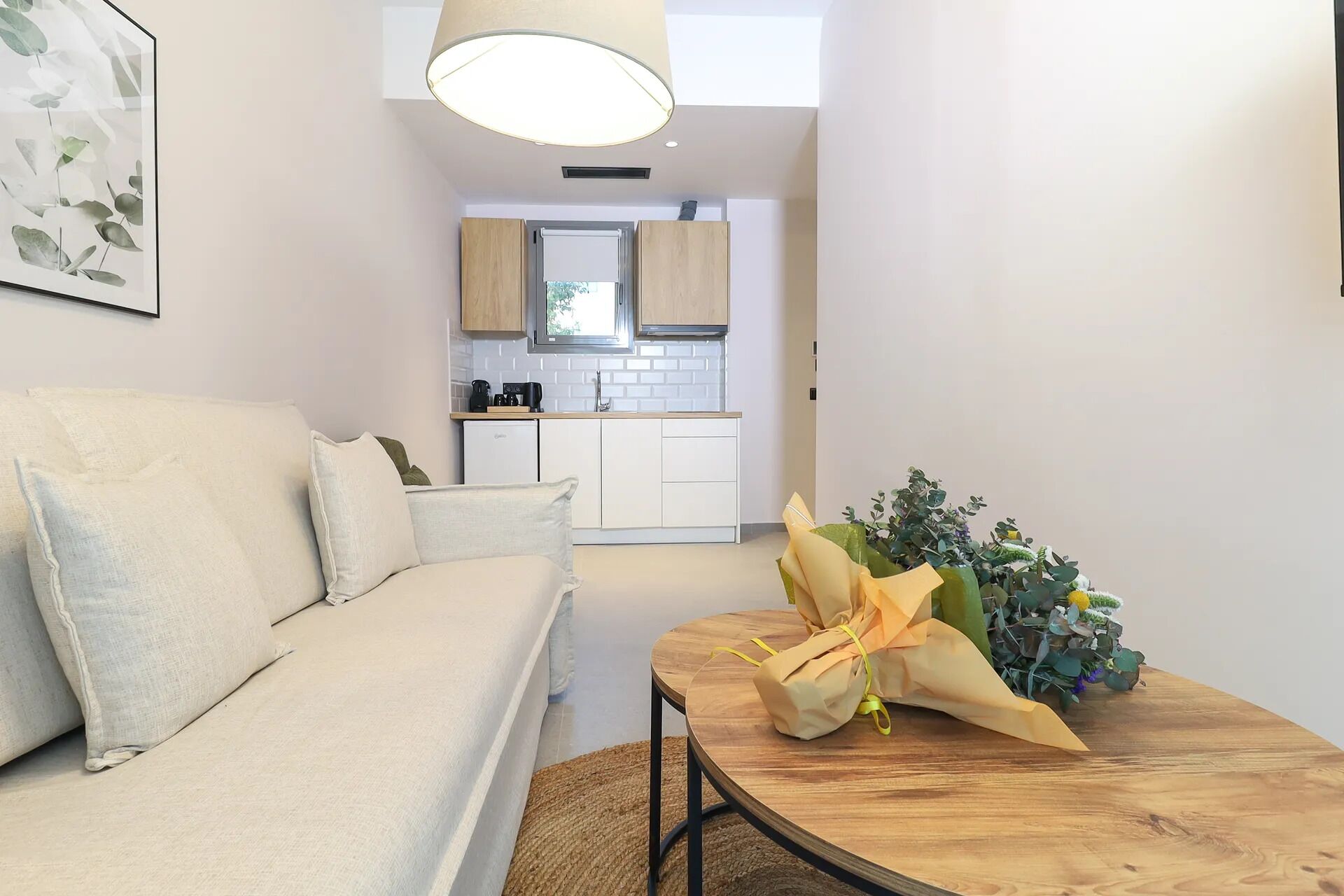 A modern living room with a sofa, a wooden coffee table with a bouquet, and a kitchen in the background.