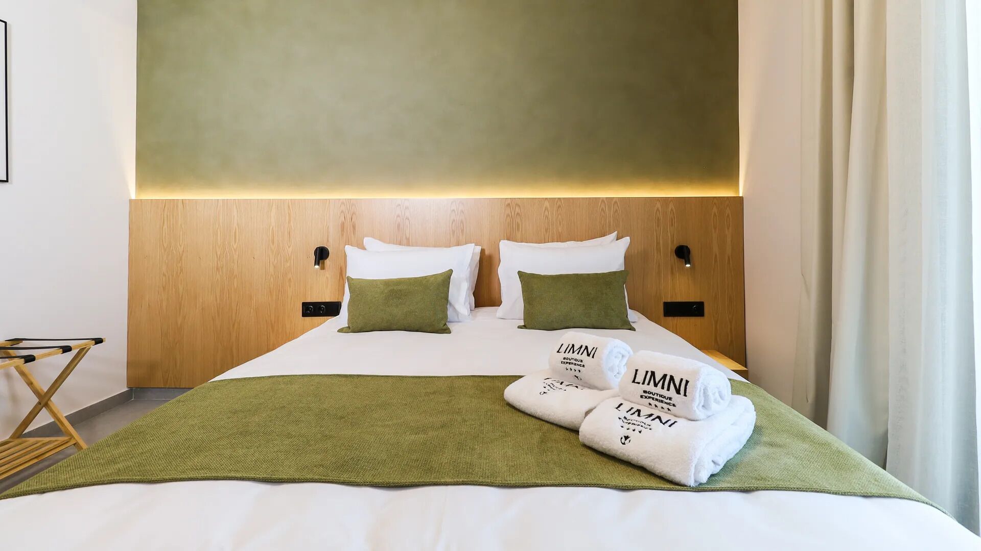 Un lit bien fait, avec du linge de lit blanc, deux oreillers verts et un plaid vert. On peut voir deux serviettes pliées sur le lit, avec l’inscription « LIMNI Boutique Experience ».