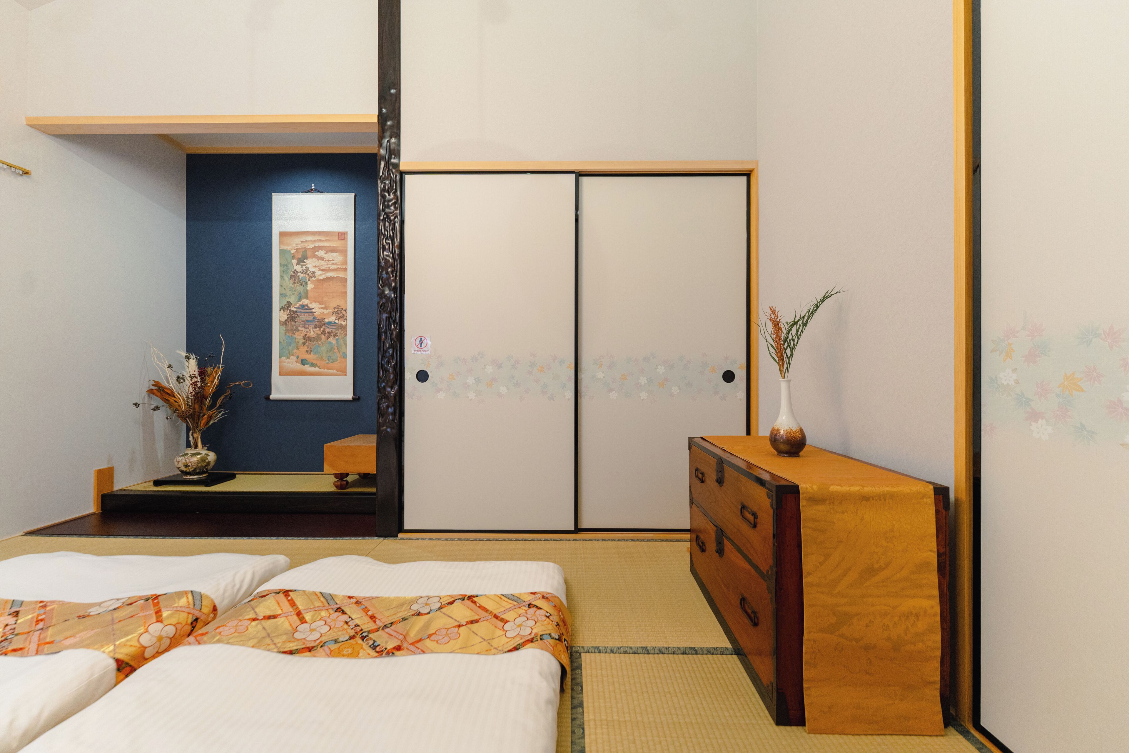 A traditional Japanese-style room with tatami flooring, a low wooden dresser, a bed with a floral bedspread, and a sliding door with floral patterns.