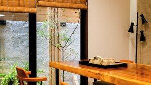A wooden dining table with chairs, a window with bamboo blinds, and a view of a stone wall and greenery outside. - Kyoto Amazing - The Wizard of Oz (Kyoto)