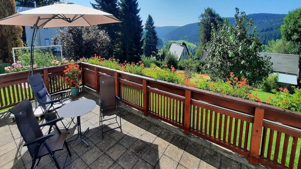 Eine Terrasse mit Tisch und Stühlen, einem Holzgeländer und Blick auf einen Garten mit Blumen und Bäumen.