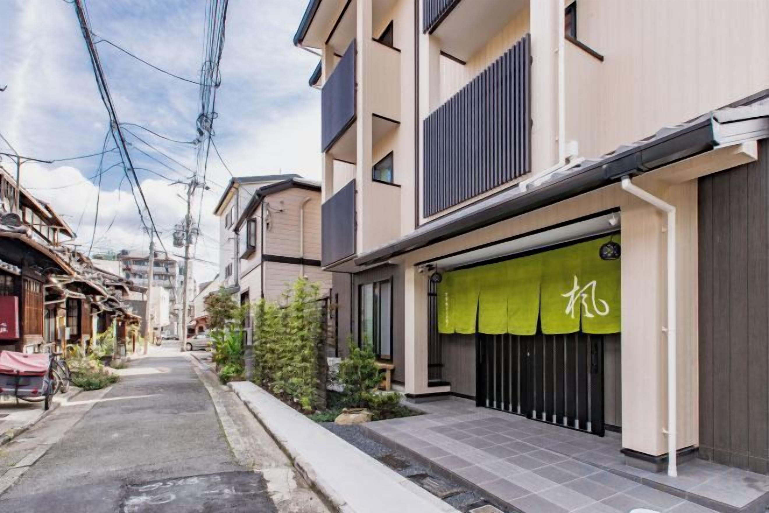 A narrow urban street with residential buildings, a green awning with Japanese characters, and overhead power lines.