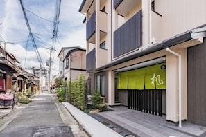 Una estrecha calle urbana con edificios residenciales, un toldo verde con caracteres japoneses y líneas eléctricas aéreas.