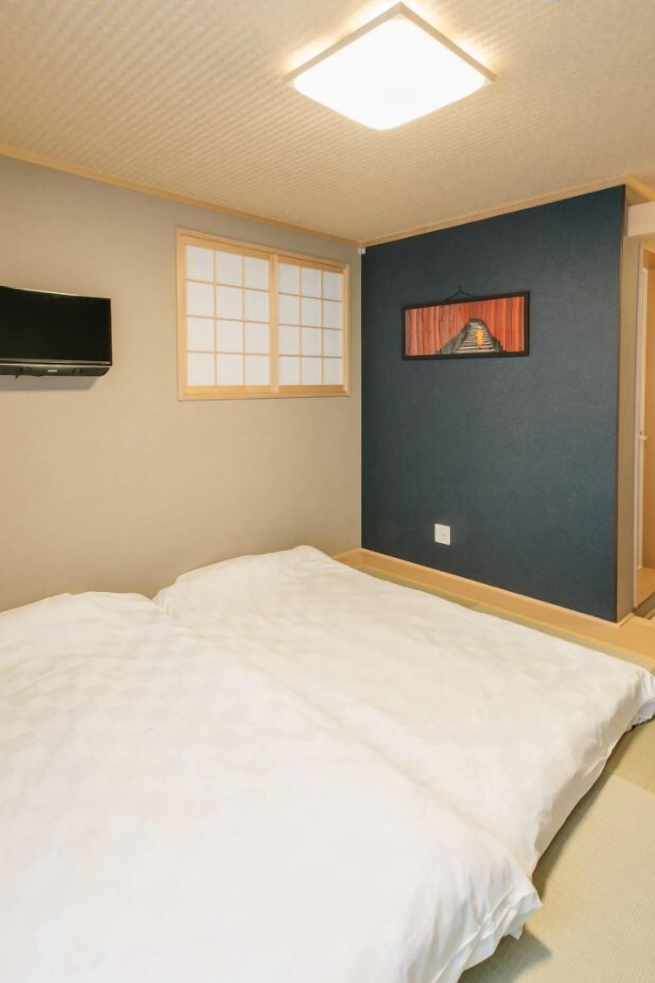 Un dormitorio con cama, televisor montado en la pared, ventana con diseño de cuadrícula y un cuadro colgado.