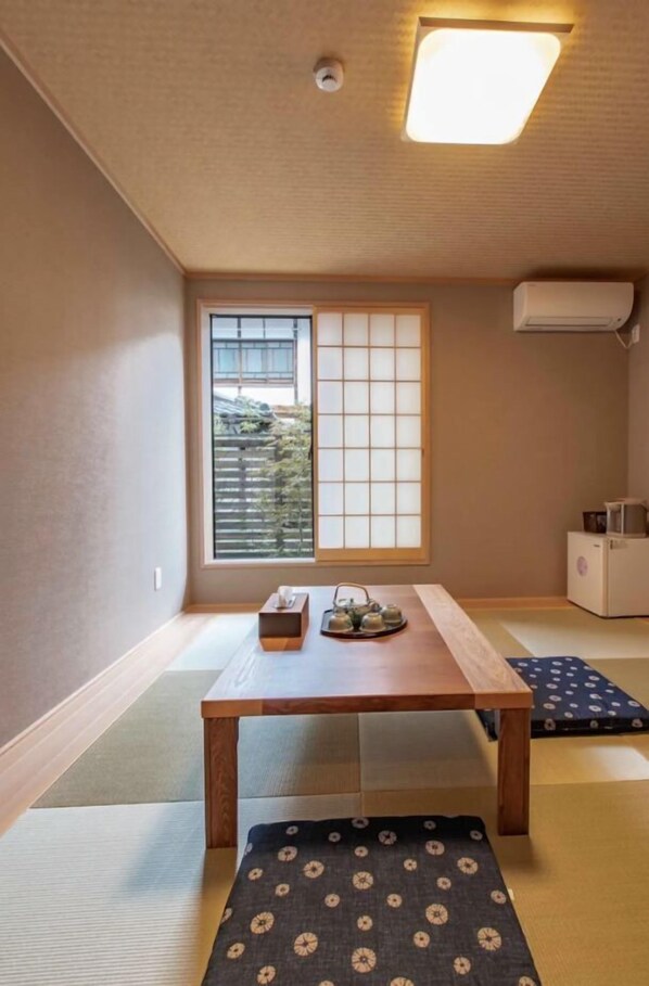 Una habitación japonesa tradicional con suelo de tatami, una mesa de madera baja, una ventana con pantalla shoji y un pequeño equipo de aire acondicionado.
