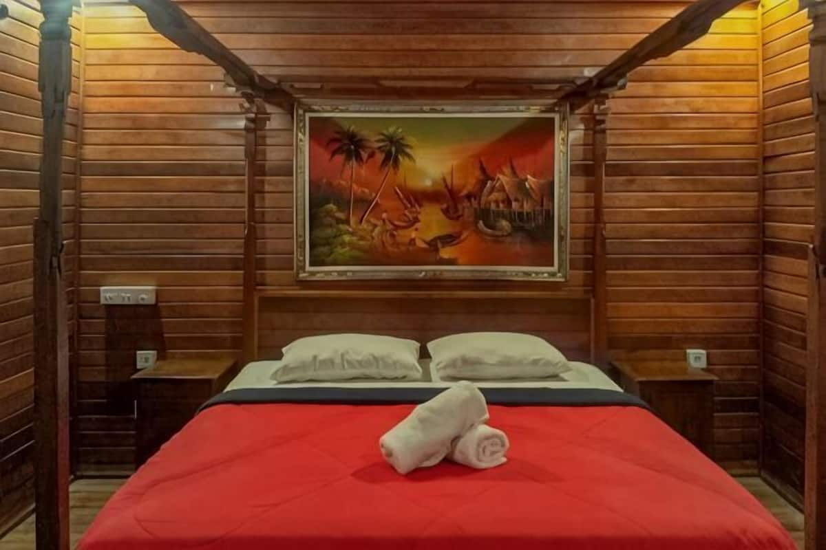 A wooden room with a bed, pillows, a painting, and a red bedspread.