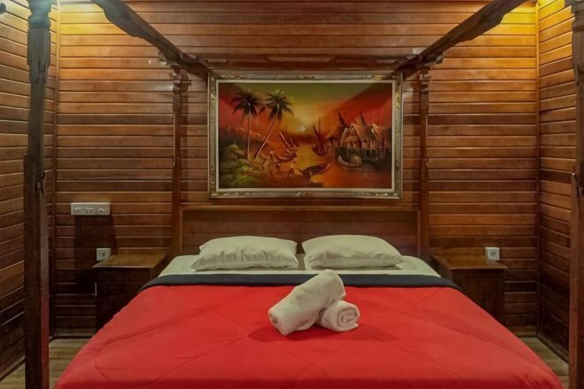 Ein Zimmer mit Holzboden, einem Bett, Kissen, einem Gemälde und einer roten Bettdecke.