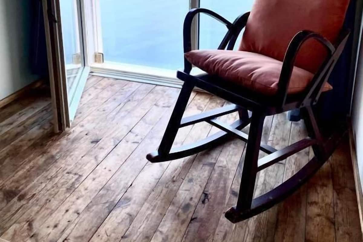 A room with a rocking chair, a wooden floor, and a large window with a view of a body of water and distant land.