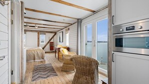 A room with a wooden ceiling, a large window with a view of the water, wicker furniture, and a built-in oven. - Sveggvika Guesthouse (Averoy)