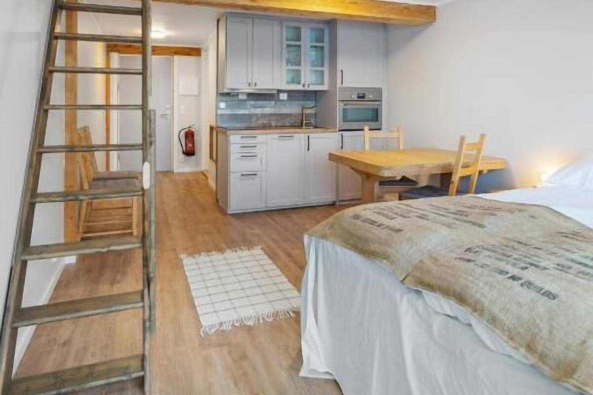 A loft-style bedroom with a wooden staircase, a kitchenette, and a dining area.