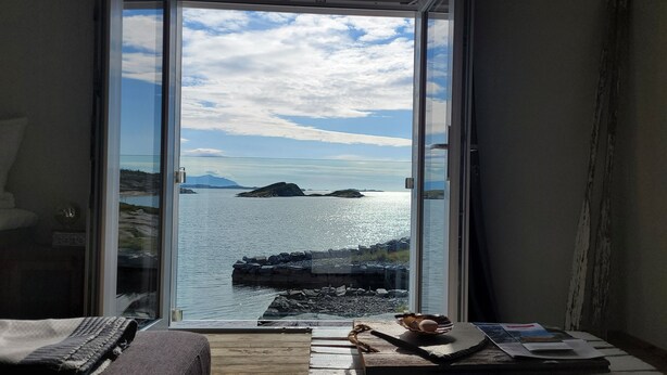 A room with a large sliding glass door leading to a scenic view of a lake and mountains. - Sveggvika Guesthouse (Averoy)
