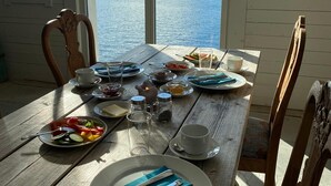 A wooden dining table with plates of food, cups, and cutlery, set against a backdrop of a lake and mountains. - Sveggvika Guesthouse (Averoy)