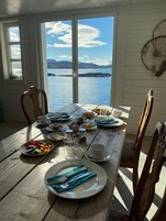 Una mesa de madera para comer con platos de comida, vasos y cubiertos, con un lago y montañas de fondo.