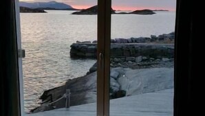 A room with a view of a rocky coastline at sunset. There is a window with curtains, a small table with a glass and a cup, and two wicker chairs. - Sveggvika Guesthouse (Averoy)
