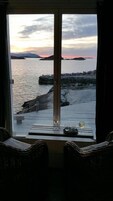 A room with a view of a rocky coastline at sunset. There is a window with curtains, a small table with a glass and a cup, and two wicker chairs.