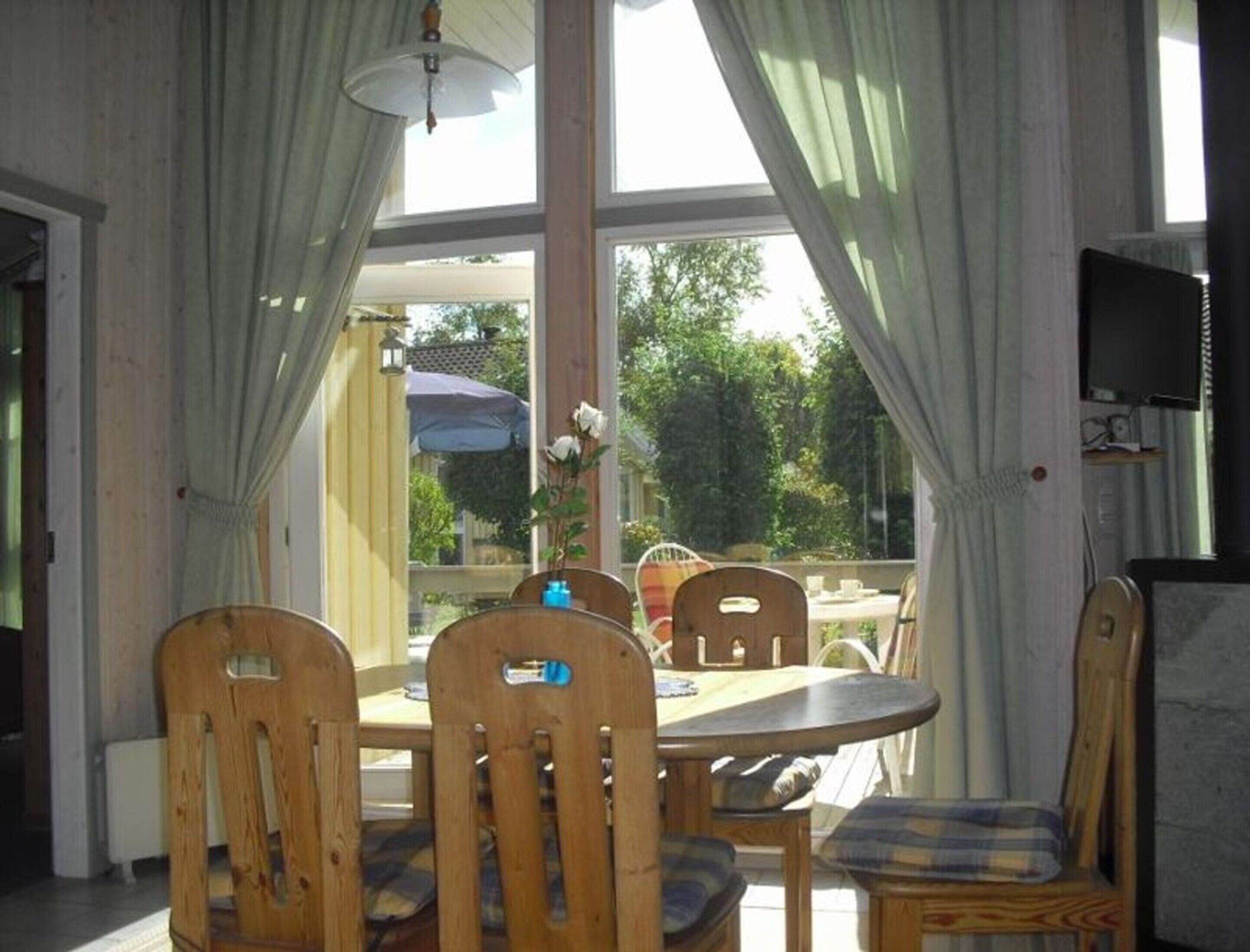 A dining area with a round table and wooden chairs, large windows with curtains, and a view of the outdoors.