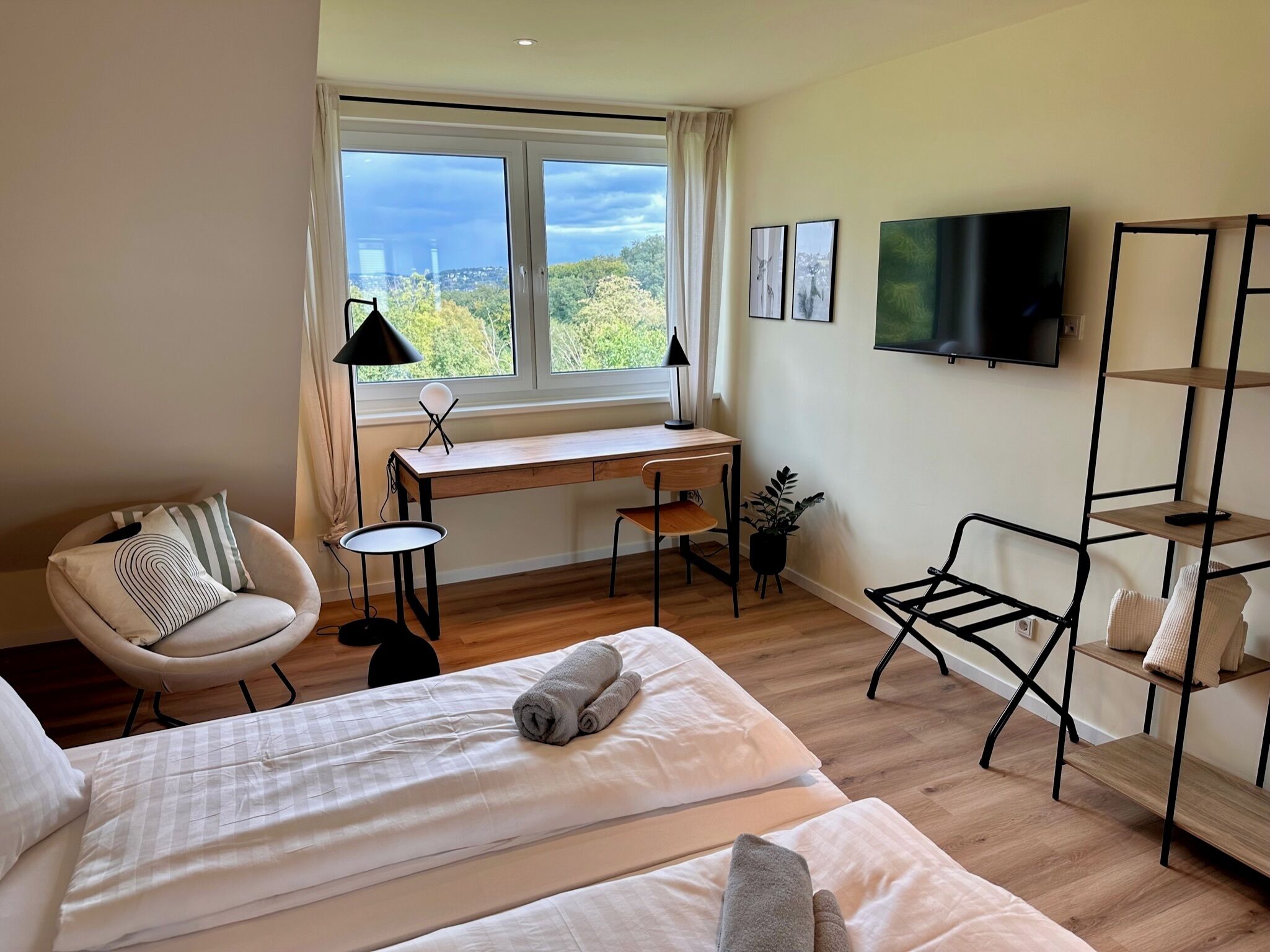 Ett hotellrum med en säng, ett skrivbord, en stol, en TV och ett fönster med utsikt över träd och byggnader.