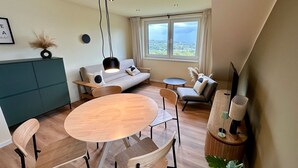 A modern living room with a sofa, a round dining table, and chairs. There is a TV stand with a TV and a plant. - Vacation apartment with lake view in Hattingen - an oasis of well-being in the middle of the Pott! (Hattingen)