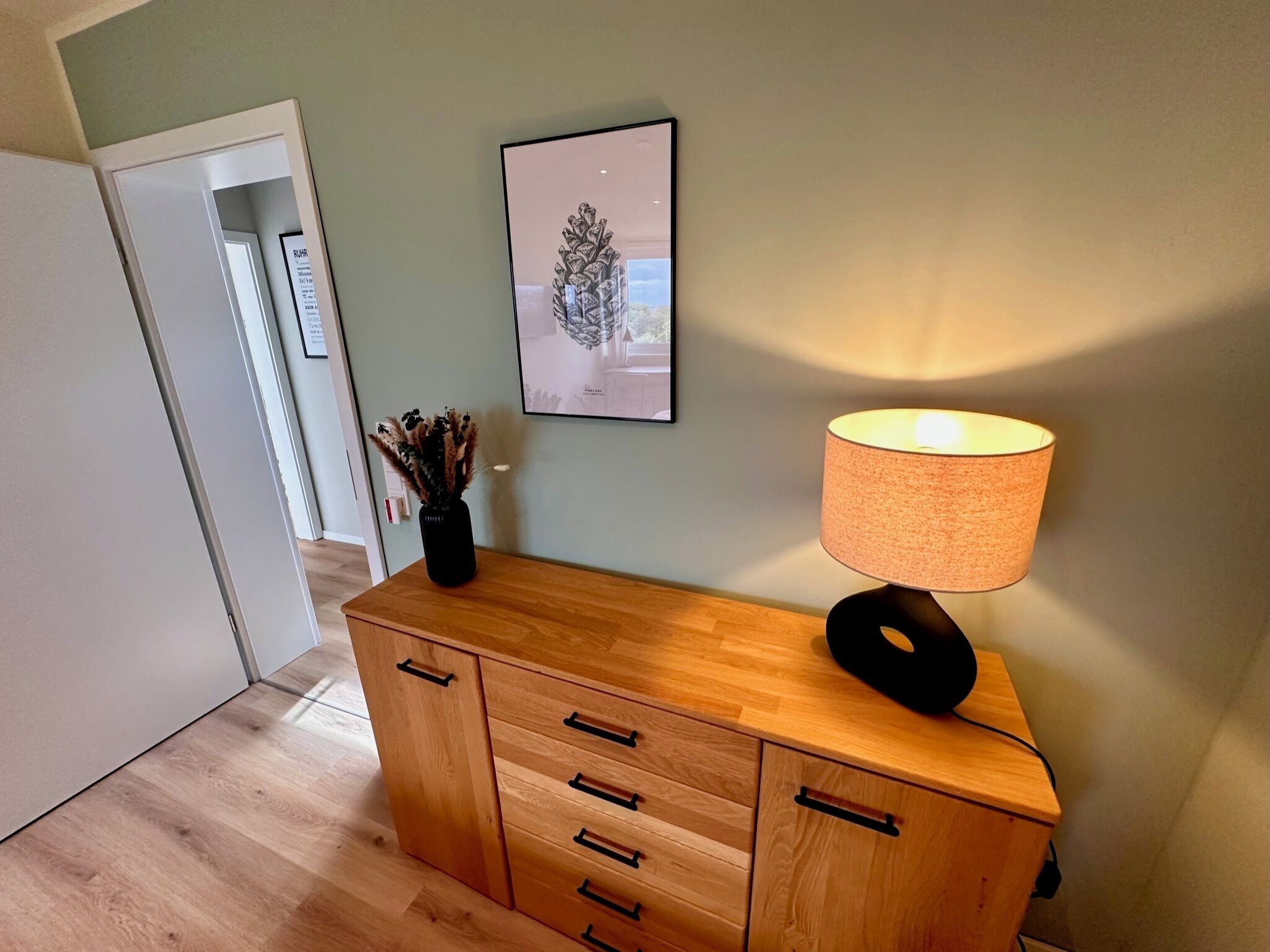 A wooden cabinet with drawers and a lamp, a framed picture on the wall, and a door in the background.