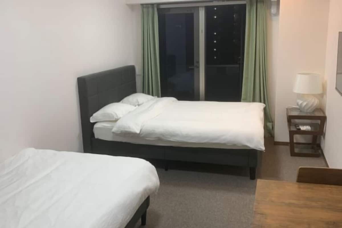 A hotel room with two beds, a small table, a lamp, and a view of the outside through a window with curtains.