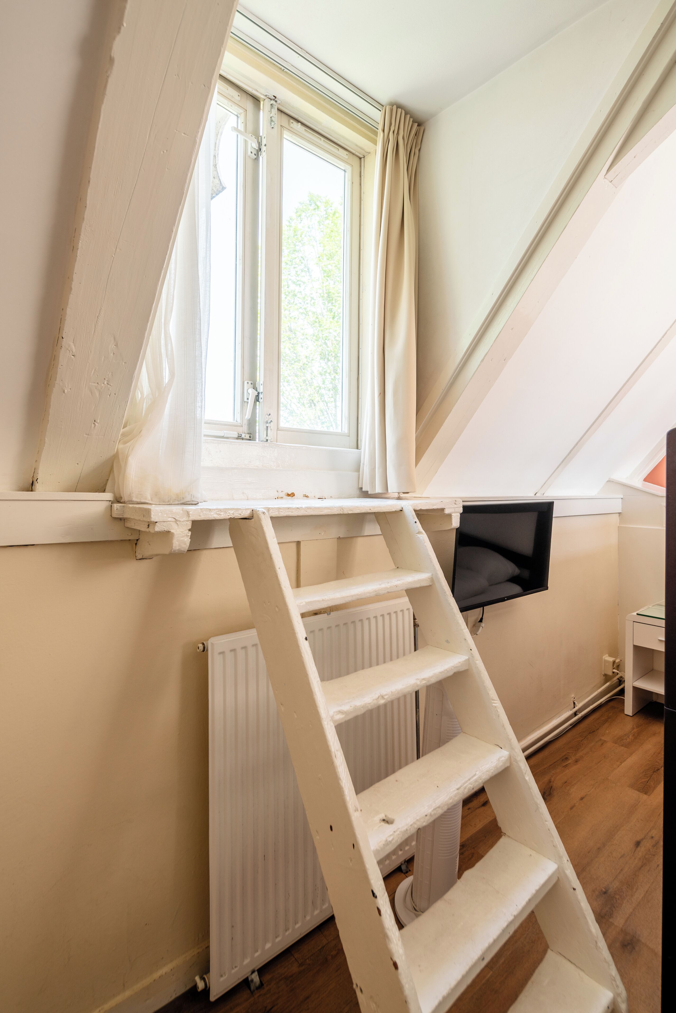 A room with a sloped ceiling, a window with curtains, a white radiator, and a white ladder.