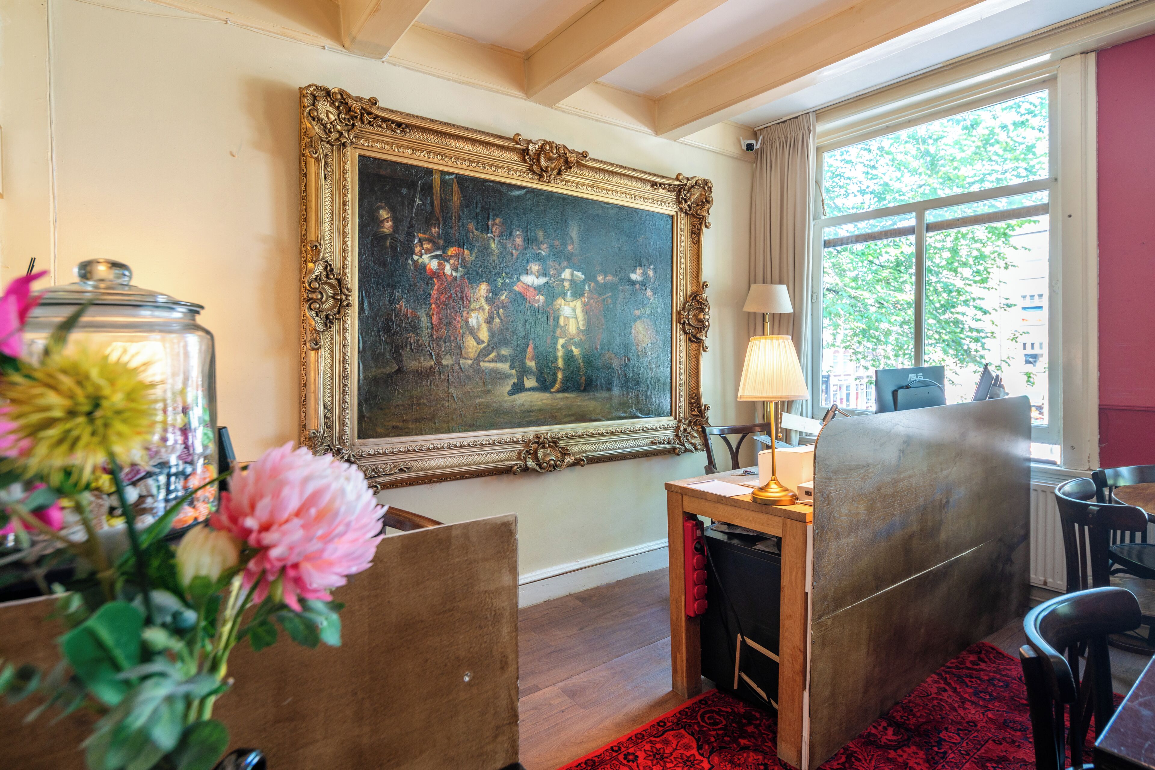 A room with a large ornate painting on the wall, a desk with a lamp, and a window with a view of trees.