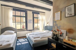A hotel room with two beds, a window with a view of buildings, a 'No Smoking' sign, a framed welcome sign, and a vase with flowers. - Hotel Prinsenhof Amsterdam (Amsterdam)