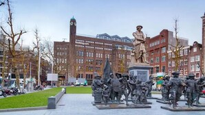 Una plaza pública con una estatua de una figura histórica y un edificio con una torre de reloj al fondo.