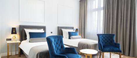 Una habitación de hotel moderna con dos camas, una zona de estar con sillas azules, una mesita redonda y una lámpara de techo.