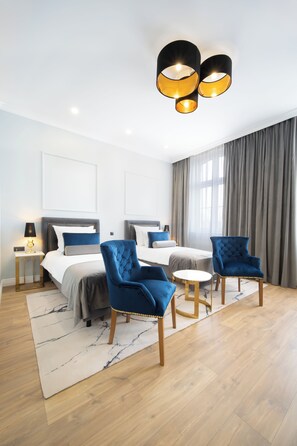 Una habitación de hotel moderna con dos camas, una zona de estar con sillas azules, una mesita redonda y una lámpara de techo.