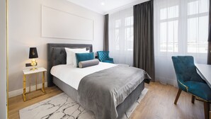 Un dormitorio moderno con una cama grande, una mesita de noche con lámpara, una silla y vistas a la ciudad a través de amplias ventanas.