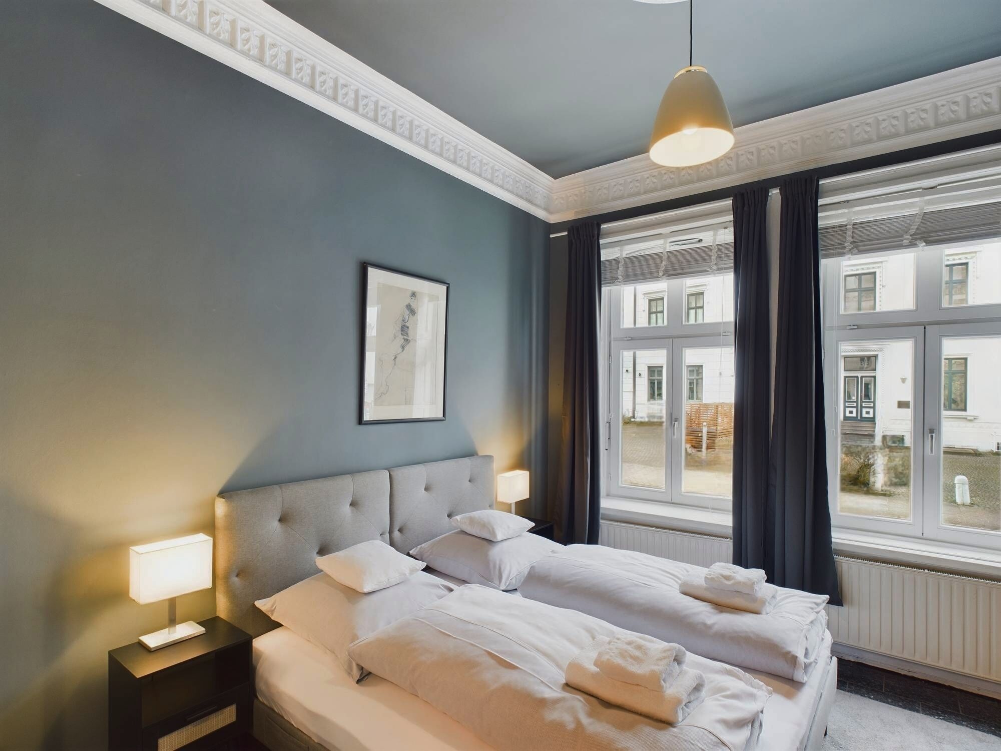 Un cuarto de hotel con una cama grande, dos mesitas de noche, una lámpara en el techo, una ventana con cortinas y un cuadro en la pared.