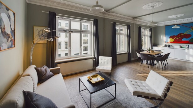 A modern living room with a sofa, armchair, and coffee table, large windows with curtains, and a dining area with chairs and a table. - Art Apartments (Hamburg)