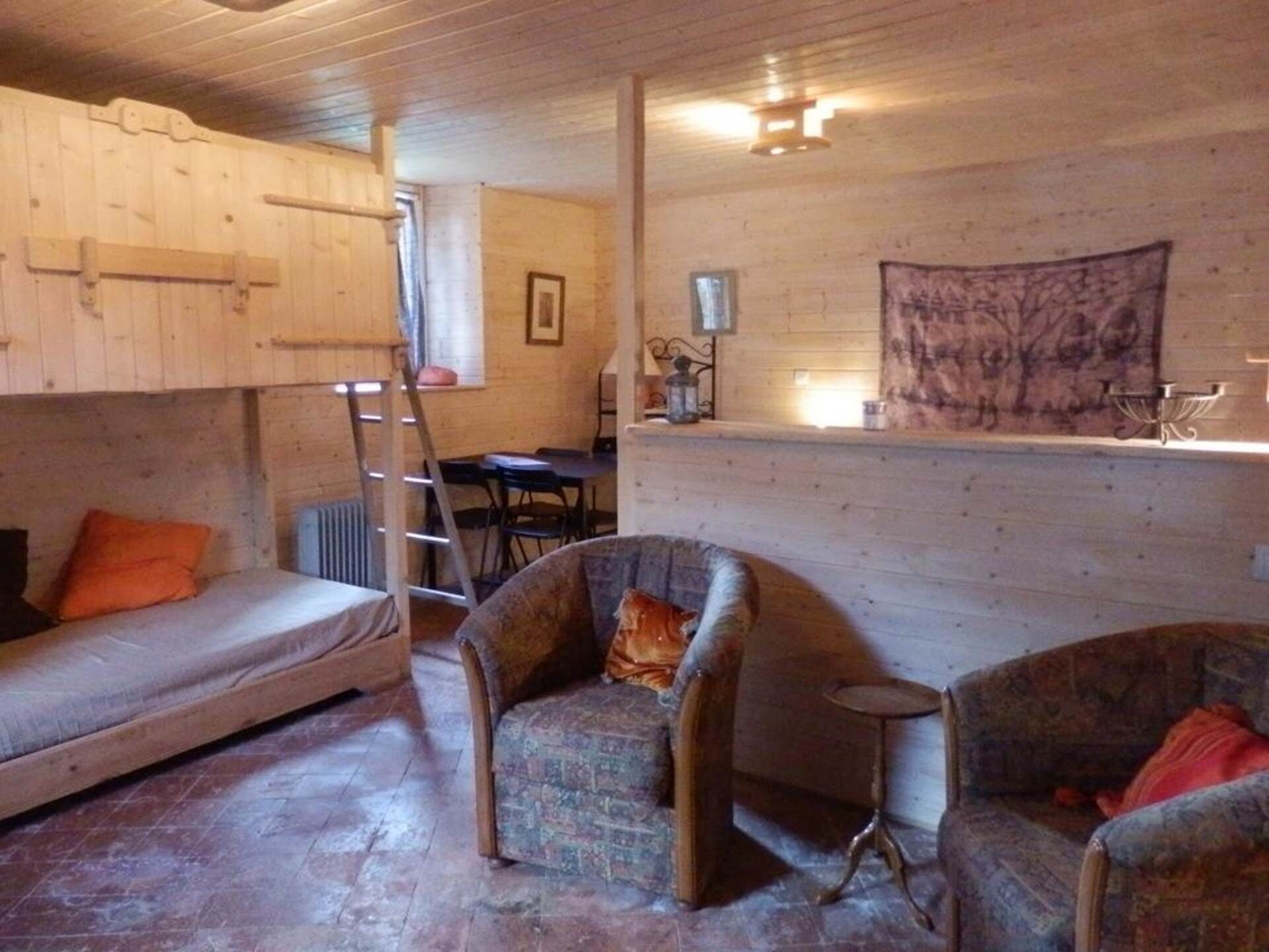 A rustic cabin interior with a bunk bed, two armchairs, a small table, and a ladder.