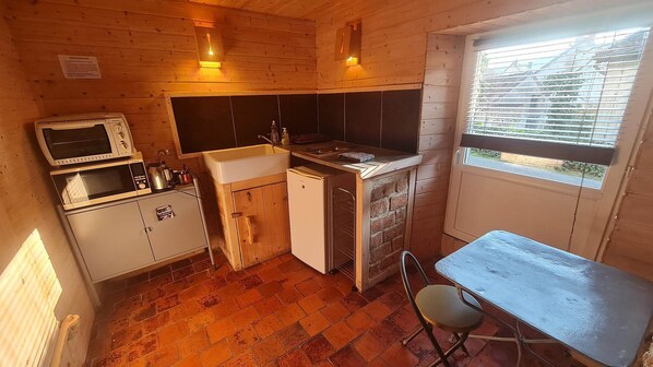 Cocina compacta con microondas, lavamanos y refrigerador pequeño. Se aprecia una ventana con persianas y una mesa de madera con una silla.