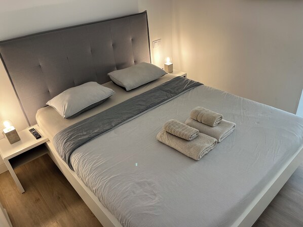 A neatly made bed with a grey and white striped bedspread, two pillows, a grey throw blanket, and two folded towels. There are bedside tables with lamps and a remote control on one of them. - LUXURY VILLA NEAR THE LAS COLINAS GOLF COURSE (Dehesa de Campoamor)