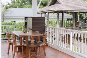 Un patio techado al aire libre con una mesa y sillas de madera, una barandilla blanca y un pabellón de jardín al fondo.