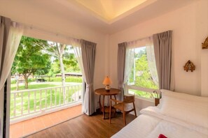 Habitación de hotel con una cama, una mesita de noche con lámpara y vistas a un jardín a través de amplios ventanales.