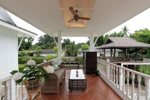 Un espacio exterior techado con ventilador de techo, mobiliario de mimbre y vistas a un jardín y una casa.