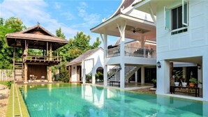 A large swimming pool with a wooden deck and a gazebo, surrounded by white buildings with balconies and outdoor seating. - Phi Private Villa: Luxury 4 beds Riverside & Pool: Near old townBreakfast, Laundry, Free pickup (Chiang Mai)