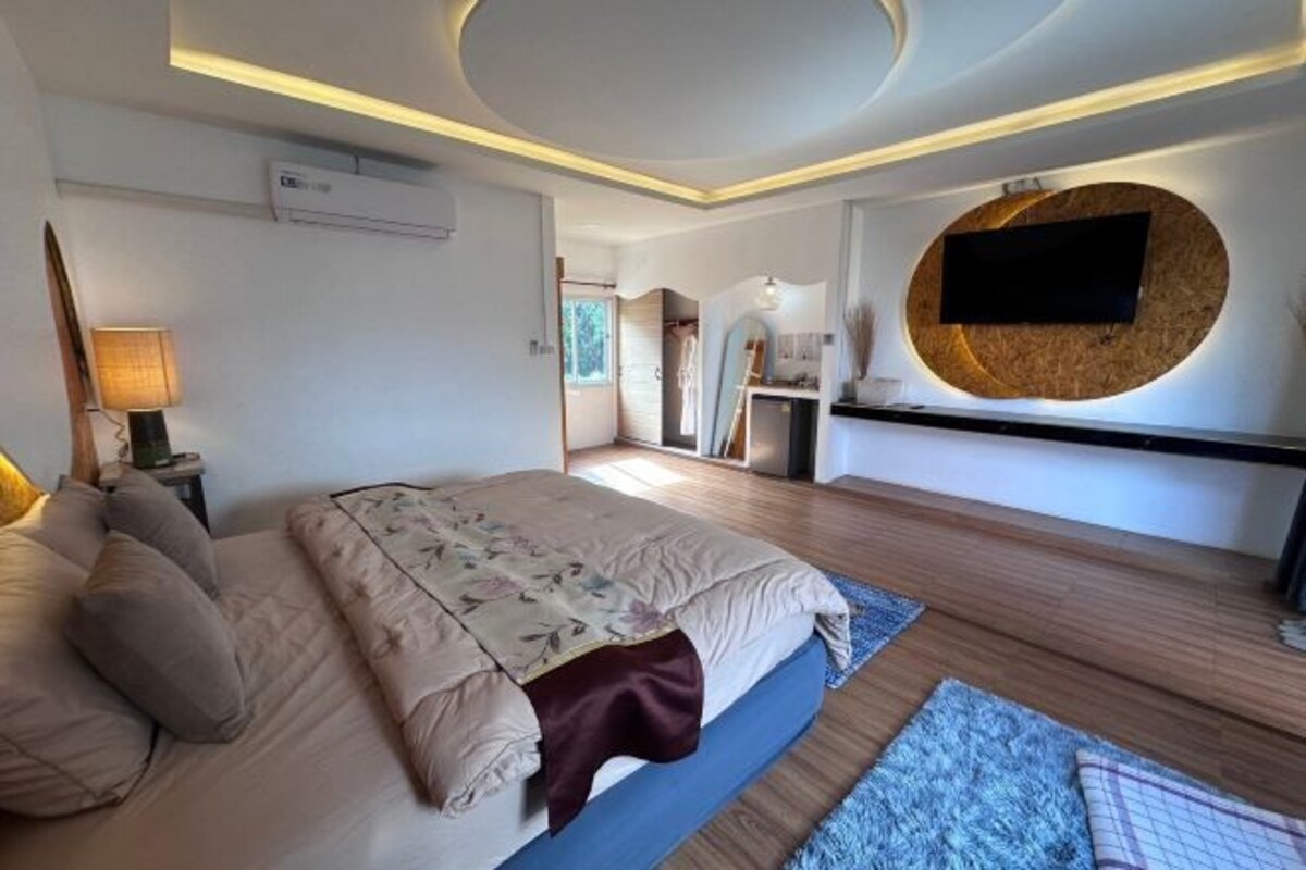 A modern bedroom with a large bed, a flat-screen TV mounted on a circular wooden panel, and a bedside table with a lamp.