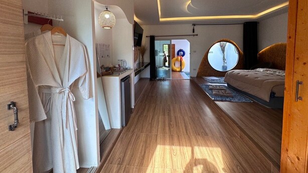 Une chambre d’hôtel moderne avec un plancher en bois, un grand lit, une fenêtre ronde et une salle de bain équipée d’un miroir et d’un porte-serviettes.