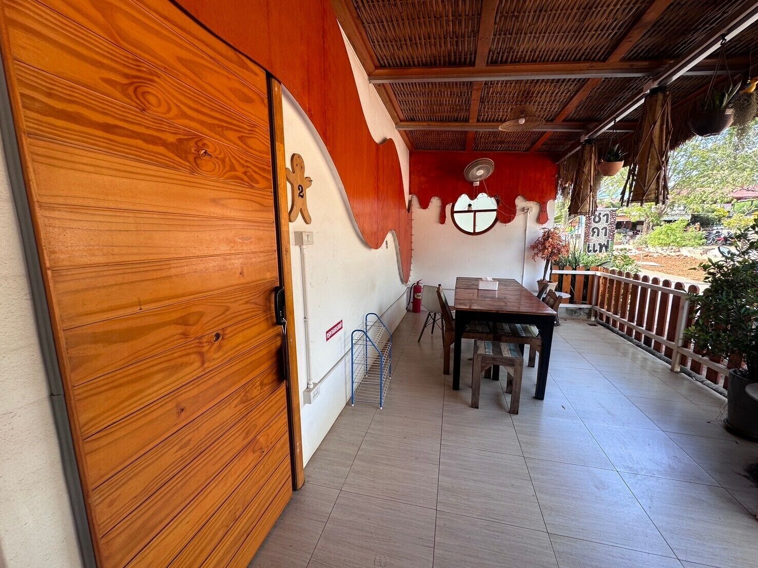 Una puerta de madera con un cartel del número 2, una mesa con sillas, un ventilador y vistas al exterior.