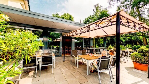 Eine Terrasse im Freien mit Tischen und Stühlen, überdacht durch ein Sonnensegel, umgeben von viel Grün.