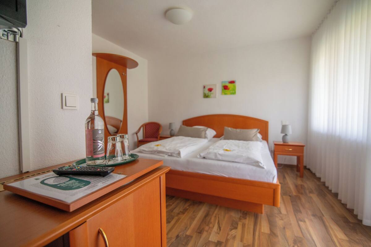 Habitación de hotel con suelo de madera, cama con ropa blanca, escritorio con control remoto, espejo y mesita de noche con lámpara.