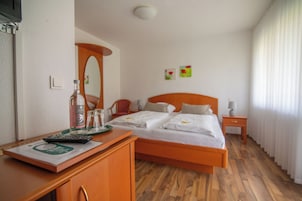 Habitación de hotel con suelo de madera, cama con ropa blanca, escritorio con control remoto, espejo y mesita de noche con lámpara.