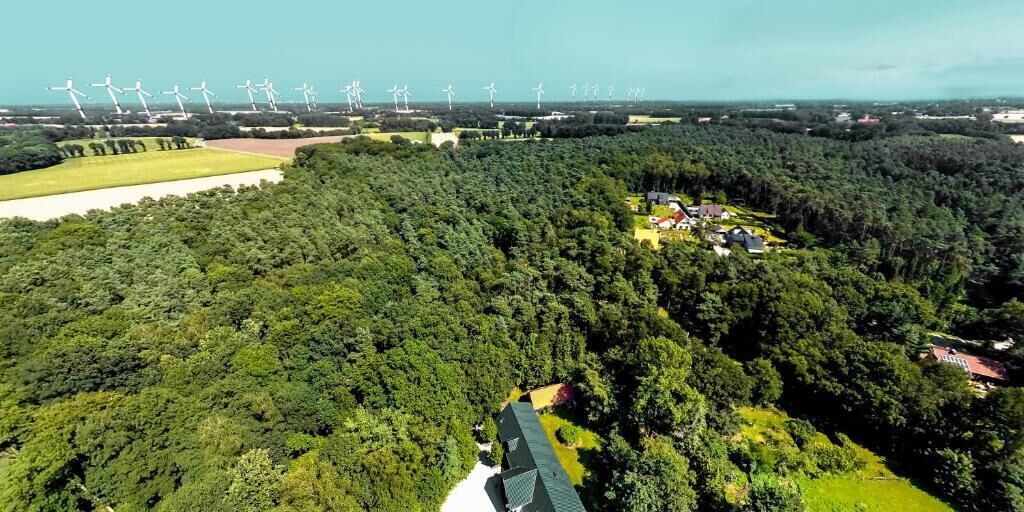 Luchtbeeld van een bebost gebied met een groep windturbines op de achtergrond en een kleine nederzetting met gebouwen en een weg.